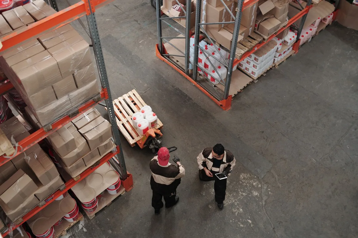 Two warehouse employees reviewing work beside storage racks, seen from above.