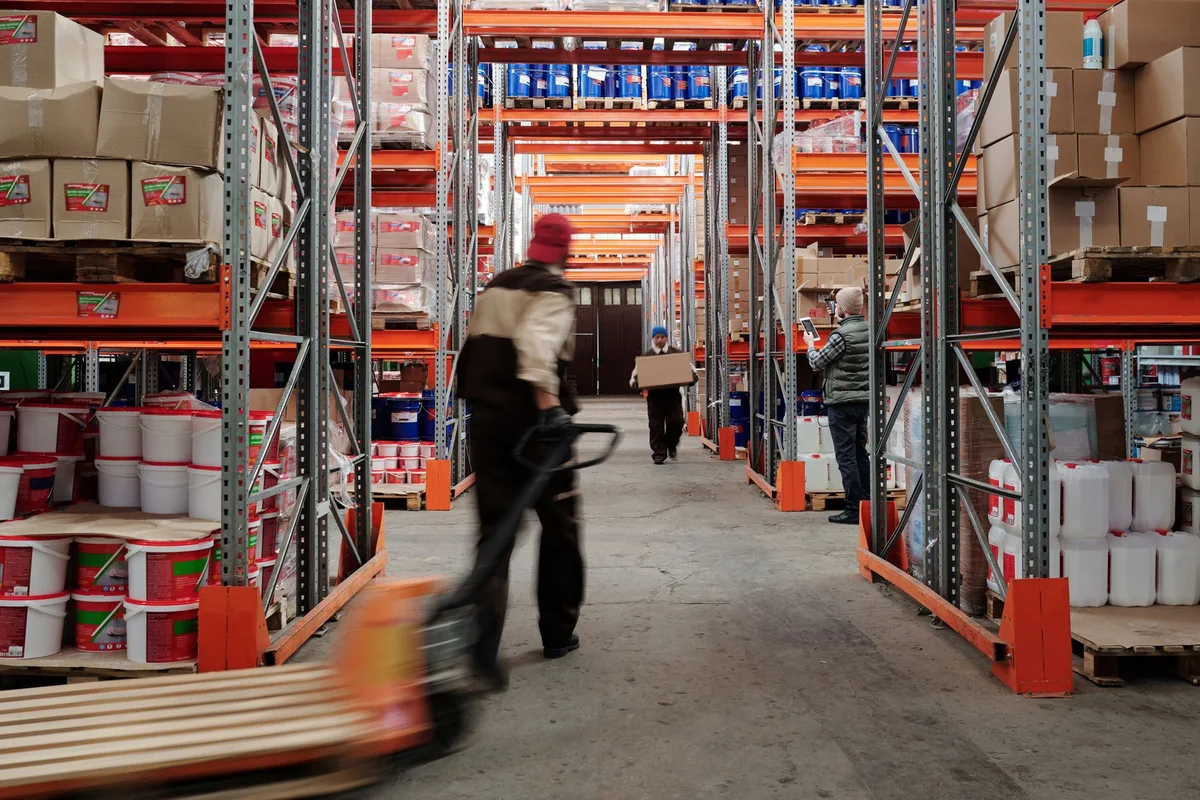 Employees moving goods through a warehouse aisle lined with storage racks.